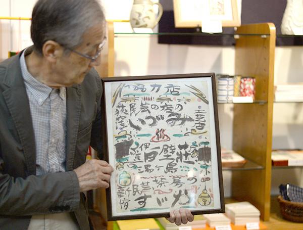 棟方志功作「民芸茶房すゞや」のポスターを持つ浩志さん。協力店として「いづみ工芸店」の名がある